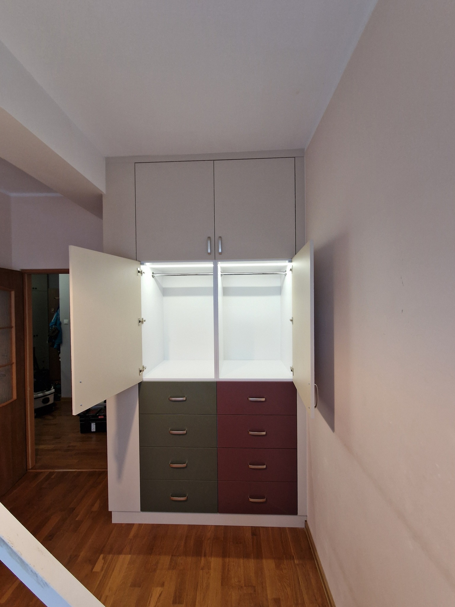 Szafa wnękowa z oświetleniem LED, otwartymi drzwiami i szufladami w odcieniach zieleni i bordo. Widok na drewnianą podłogę i fragment sąsiedniego pomieszczenia.