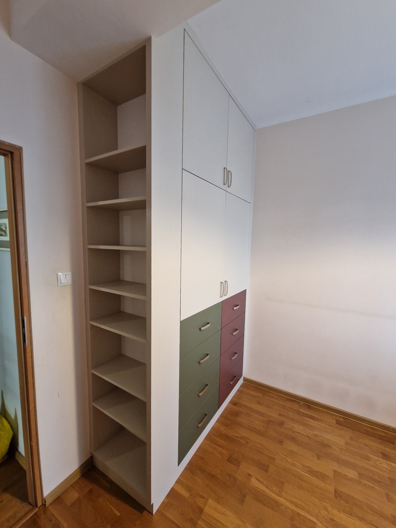 Biała szafa wnękowa z półkami i kolorowymi szufladami (zieleń, bordo) w jasnym pokoju z drewnianą podłogą. Minimalistyczne uchwyty.