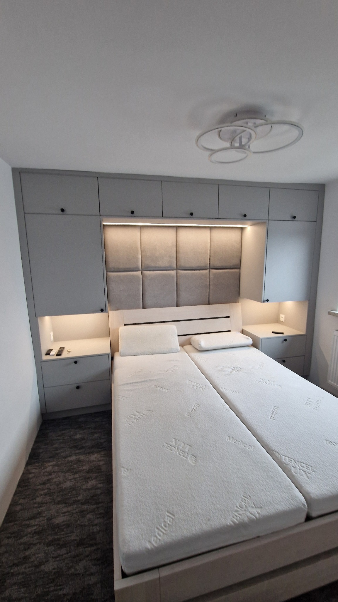 Szara zabudowa szaf wnękowych nad łóżkiem z tapicerowanym wezgłowiem i oświetleniem LED. Szafki nocne z szufladami i wnękami. Nowoczesny design, minimalistyczny styl.