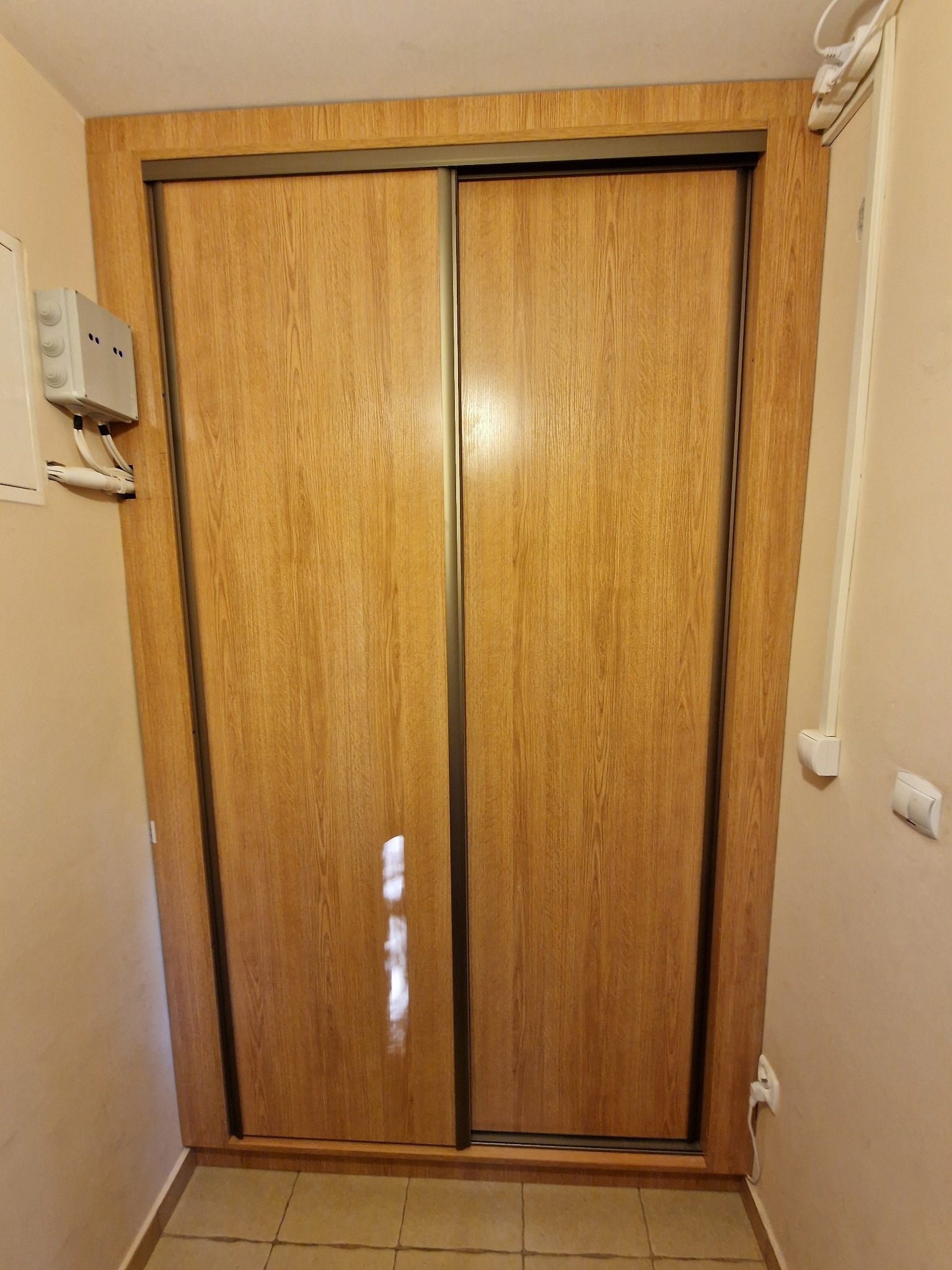 Szafa wnękowa z przesuwnymi drzwiami o drewnianej fakturze, z ciemnymi obramowaniami, umieszczona w korytarzu. Widoczna instalacja elektryczna na ścianie.