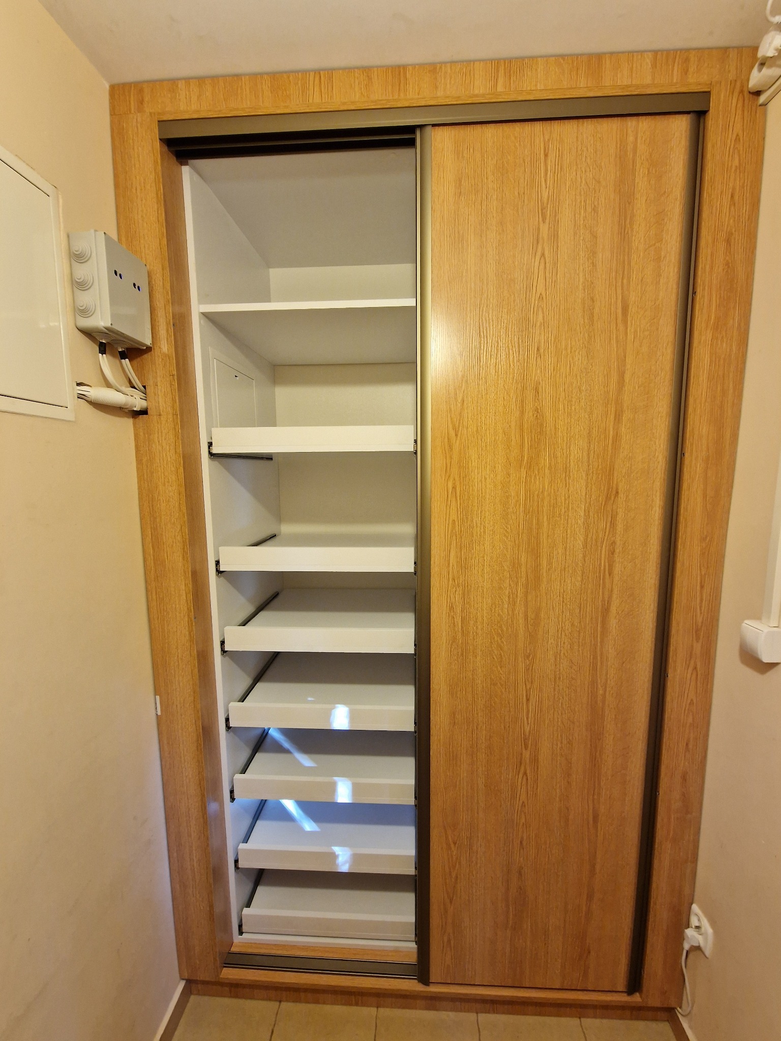 Szafa wnękowa z przesuwnymi drzwiami o drewnianej fakturze, z widocznymi półkami wewnątrz. Oświetlenie sufitowe rzuca cień na białe półki.