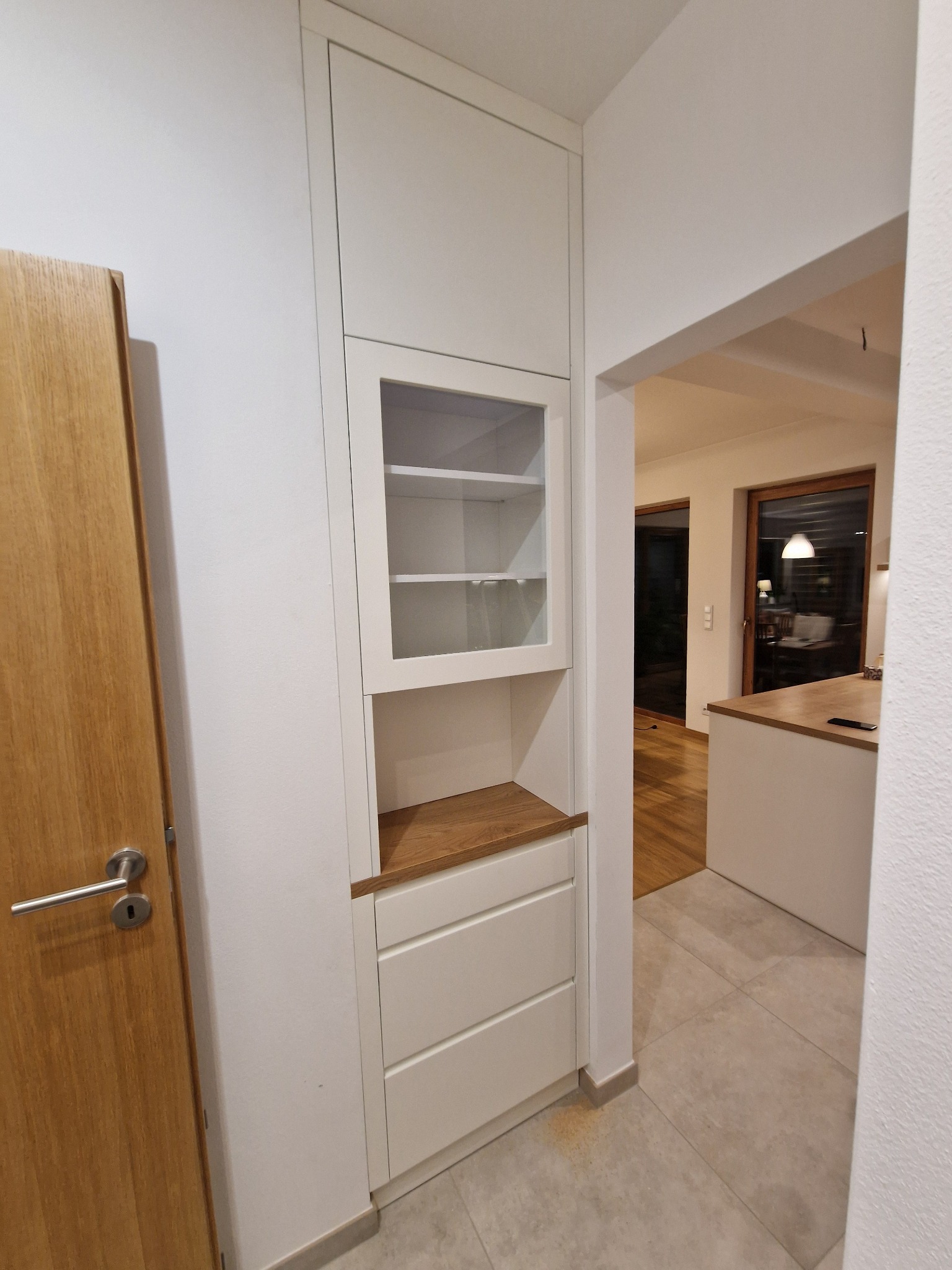 Biała szafa wnękowa z przeszkloną witryną i drewnianym blatem, wkomponowana w korytarz, widok z perspektywy wejścia do pomieszczenia. Minimalistyczny design.