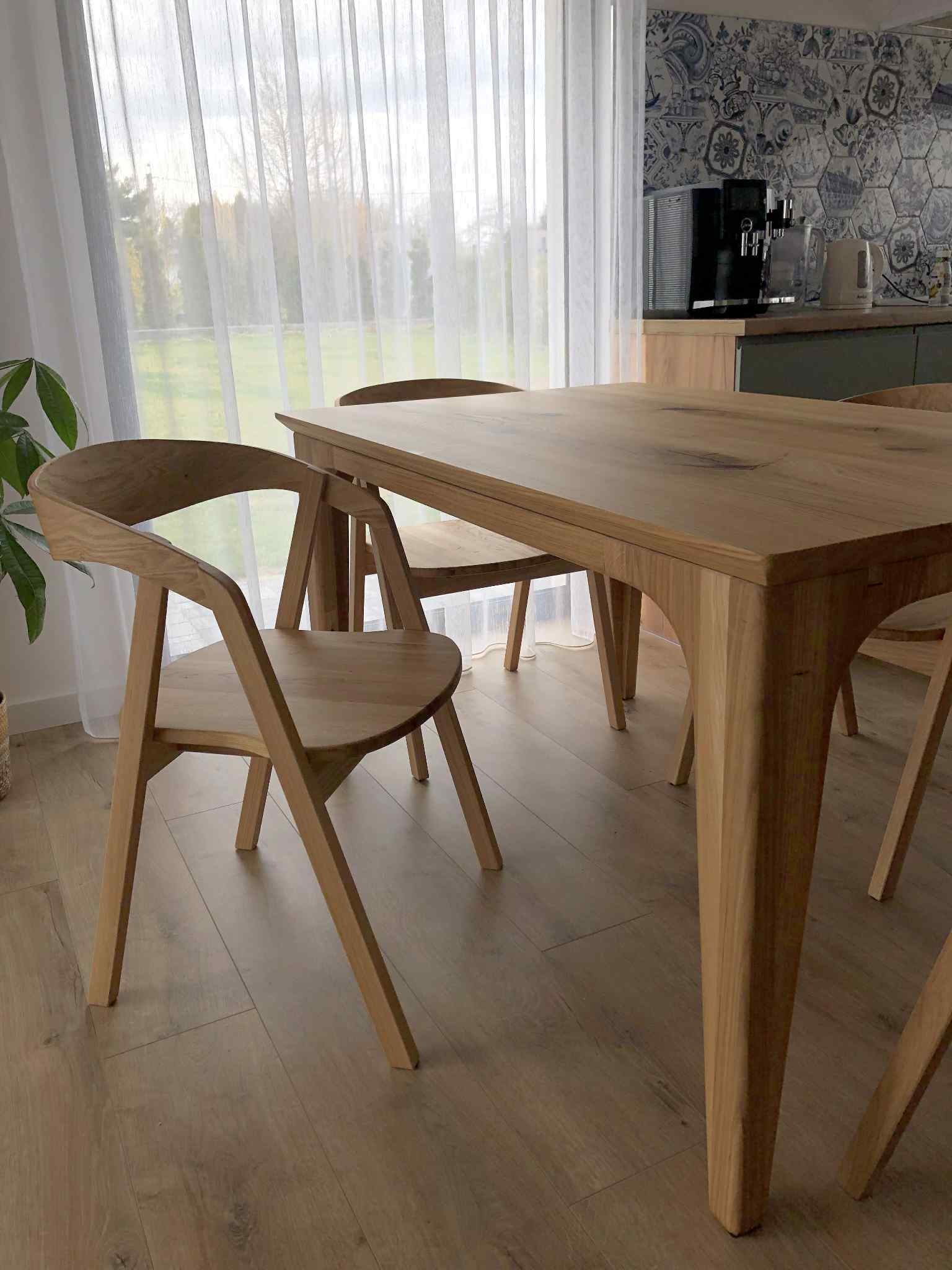 Drewniany stół z krzesłami w jasnym wnętrzu. Widoczne detale wykonania mebli i wzorzysta ściana w tle. Naturalne światło podkreśla ciepłą kolorystykę drewna.