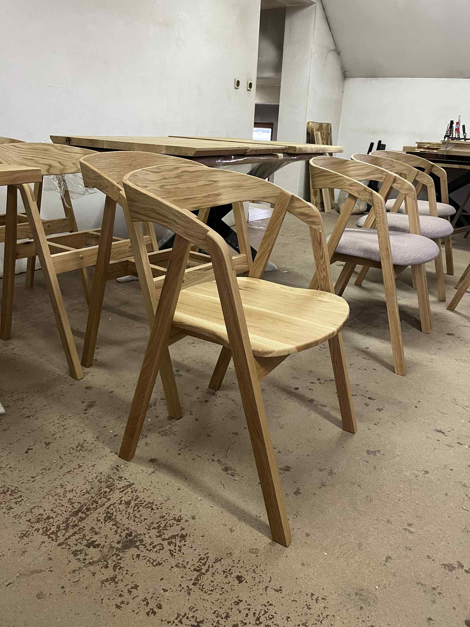 Drewniane krzesła i stół w warsztacie stolarskim. Krzesła z jasnego drewna z szarymi siedziskami. W tle widoczny stół z blatem z litego drewna i metalowymi nogami.