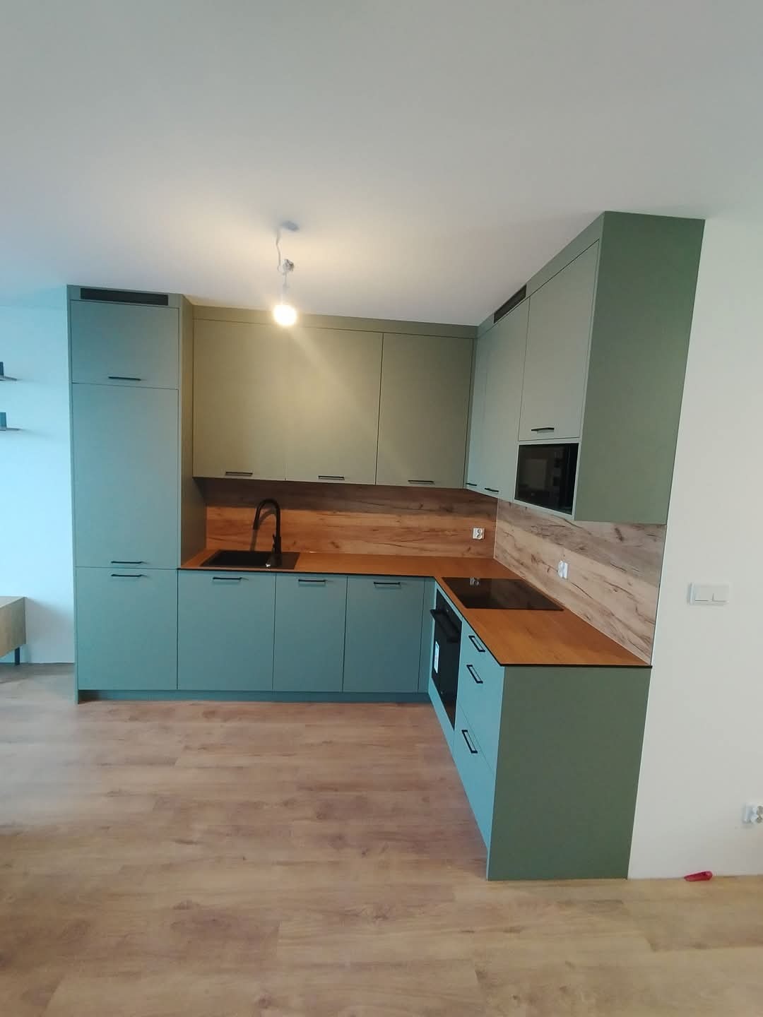 Nowoczesna kuchnia w odcieniach zieleni z drewnianym blatem i czarną armaturą. Minimalistyczny design, fronty bez uchwytów, oświetlenie punktowe. Ujęcie z perspektywy podłogi.