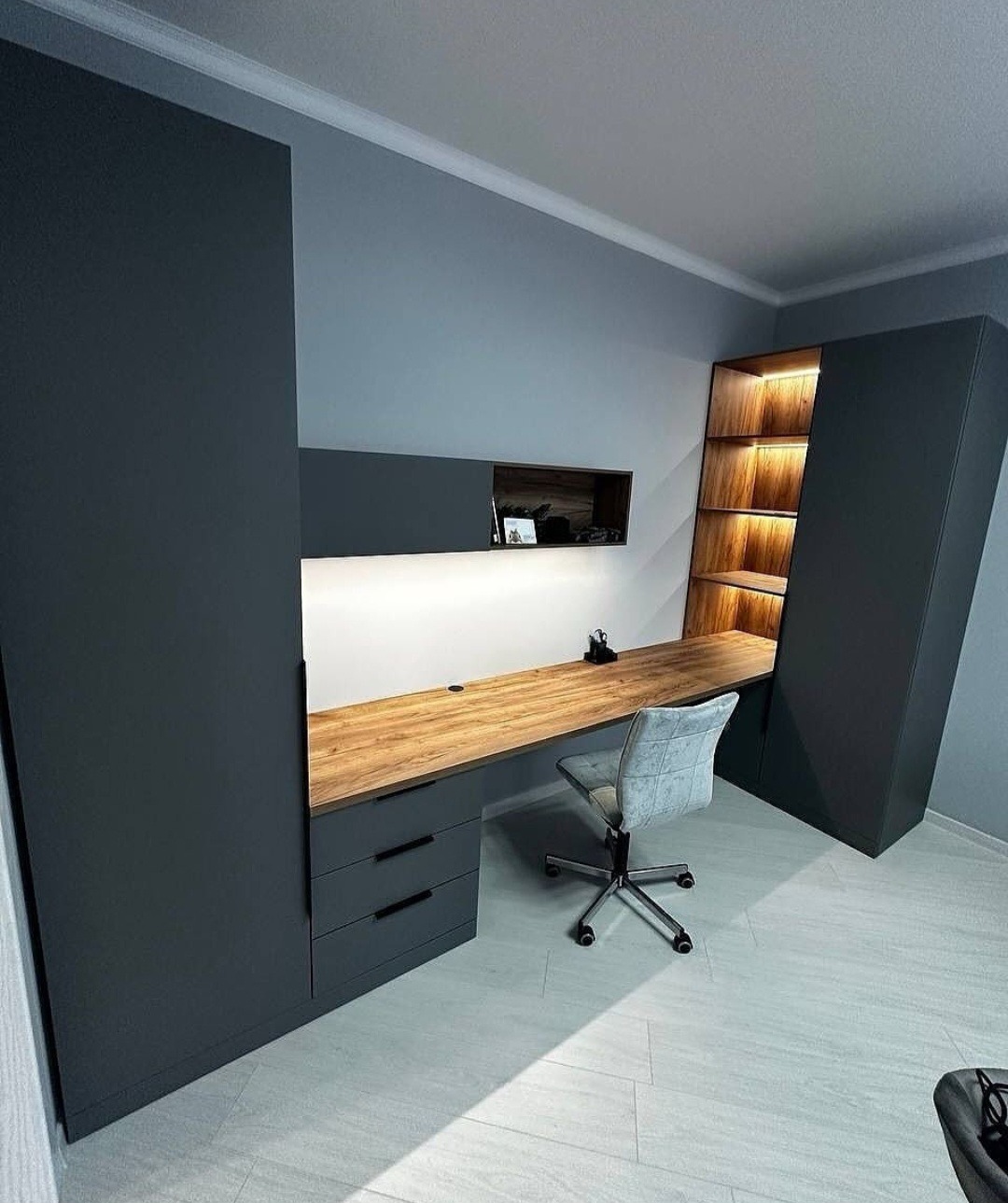 Nowoczesne biurko zintegrowane z szafą wnękową w odcieniach szarości i drewna, oświetlone półki, krzesło biurowe na kółkach, minimalistyczny design.