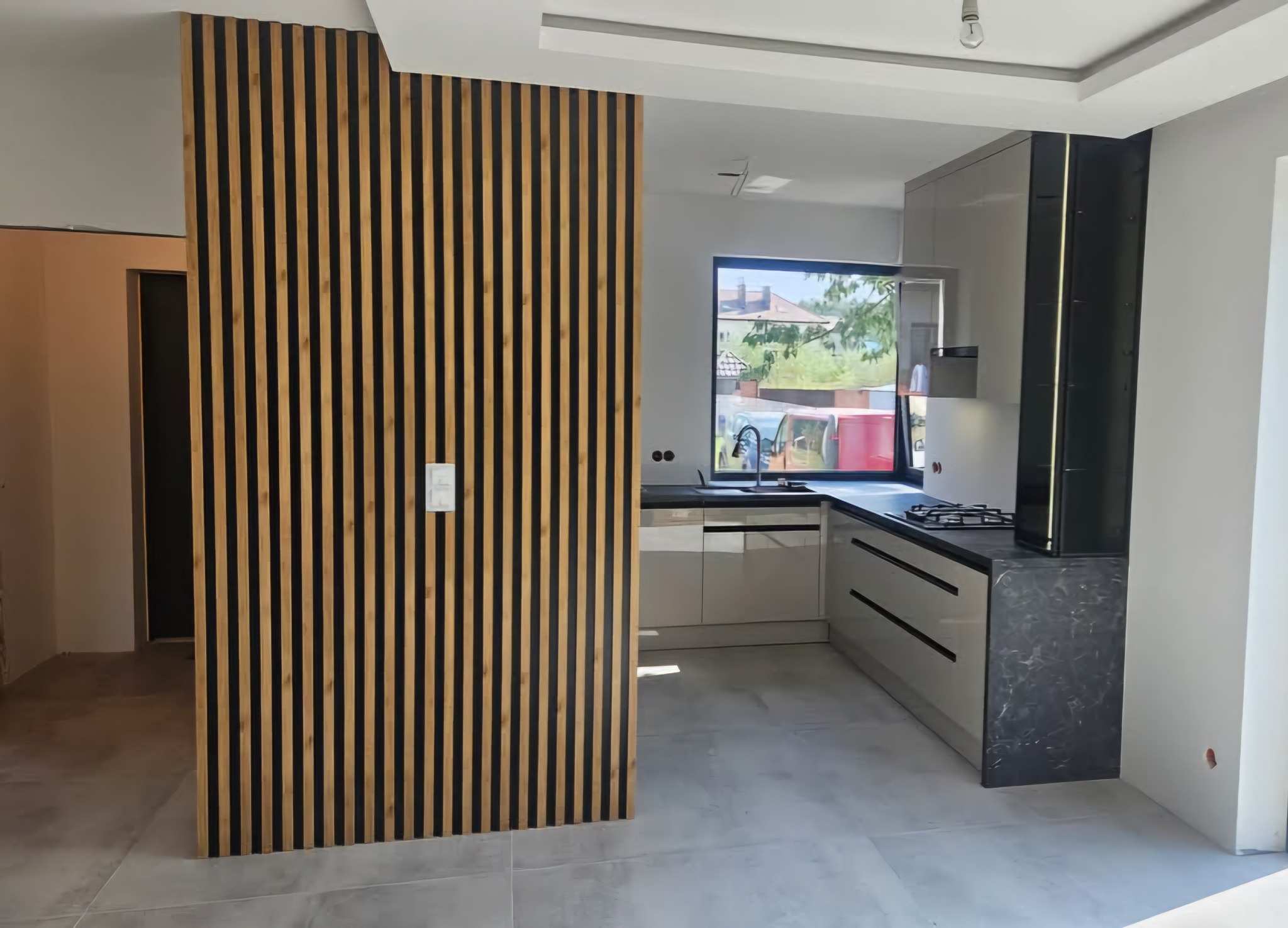 Nowoczesna kuchnia z zabudową na wymiar, szare fronty, czarny blat i drewniana ścianka działowa z lameli. Minimalistyczny design, jasne wnętrze.