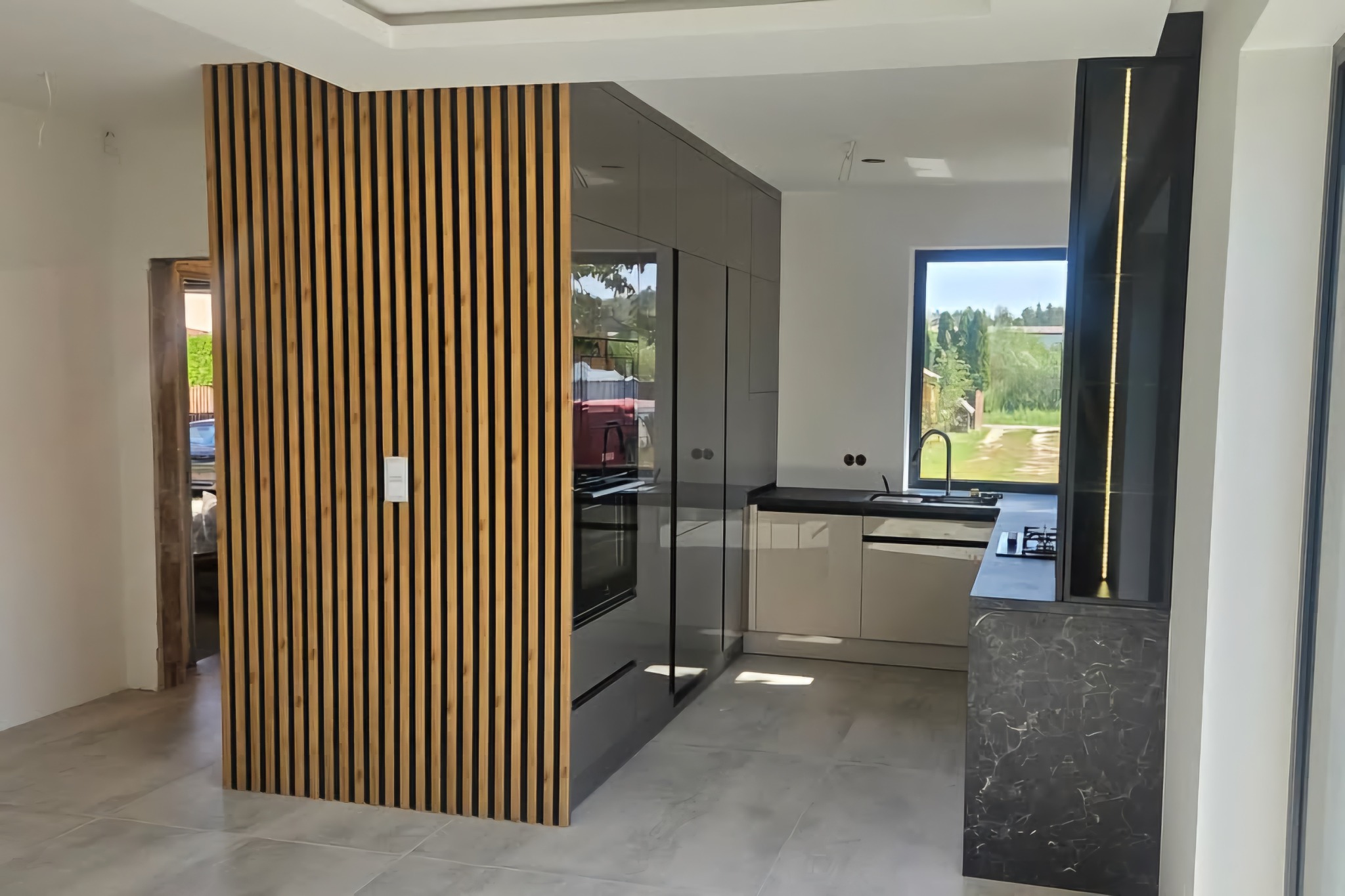 Nowoczesna kuchnia z drewnianą ścianką działową i zabudową w odcieniach szarości. Minimalistyczny design, duże okno z widokiem na zieleń.