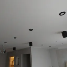 Biały sufit z zamontowanymi czarnymi, okrągłymi oprawami oświetleniowymi, widoczne ściany w szarych odcieniach w trakcie wykańczania, surowe wykończenie ścian