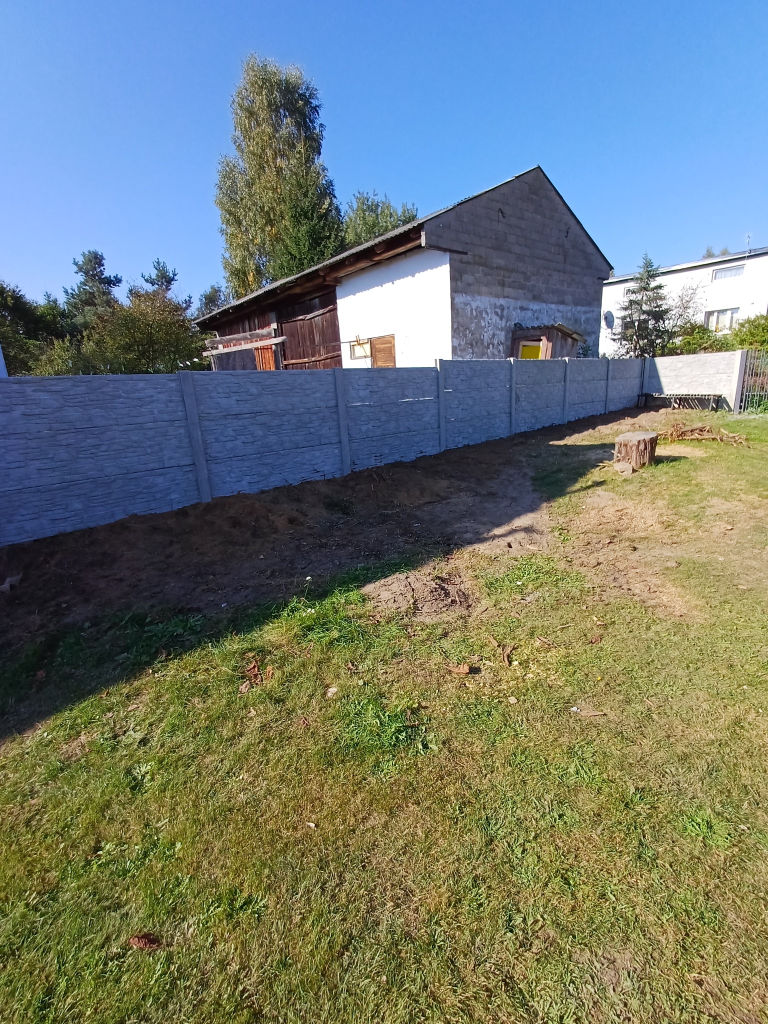 Długi, szary mur z betonowych prefabrykatów oddziela posesję z trawnikiem od sąsiedniej działki zabudowanej budynkami gospodarczymi i domami jednorodzinnymi.