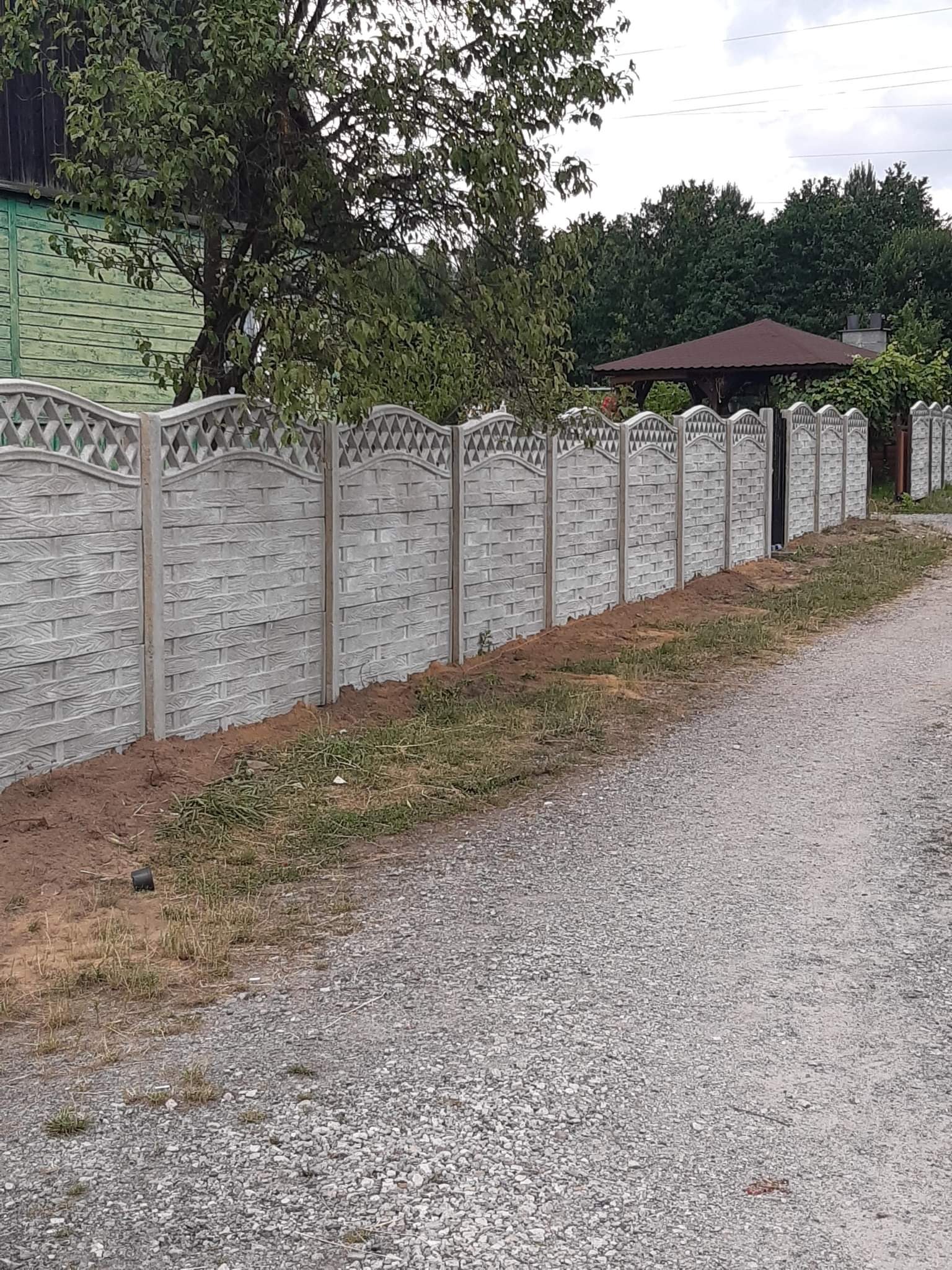 Długi, szary, betonowy płot z dekoracyjnym wzorem na szczycie, oddzielający posesję od żwirowej drogi, z widoczną altaną w tle.