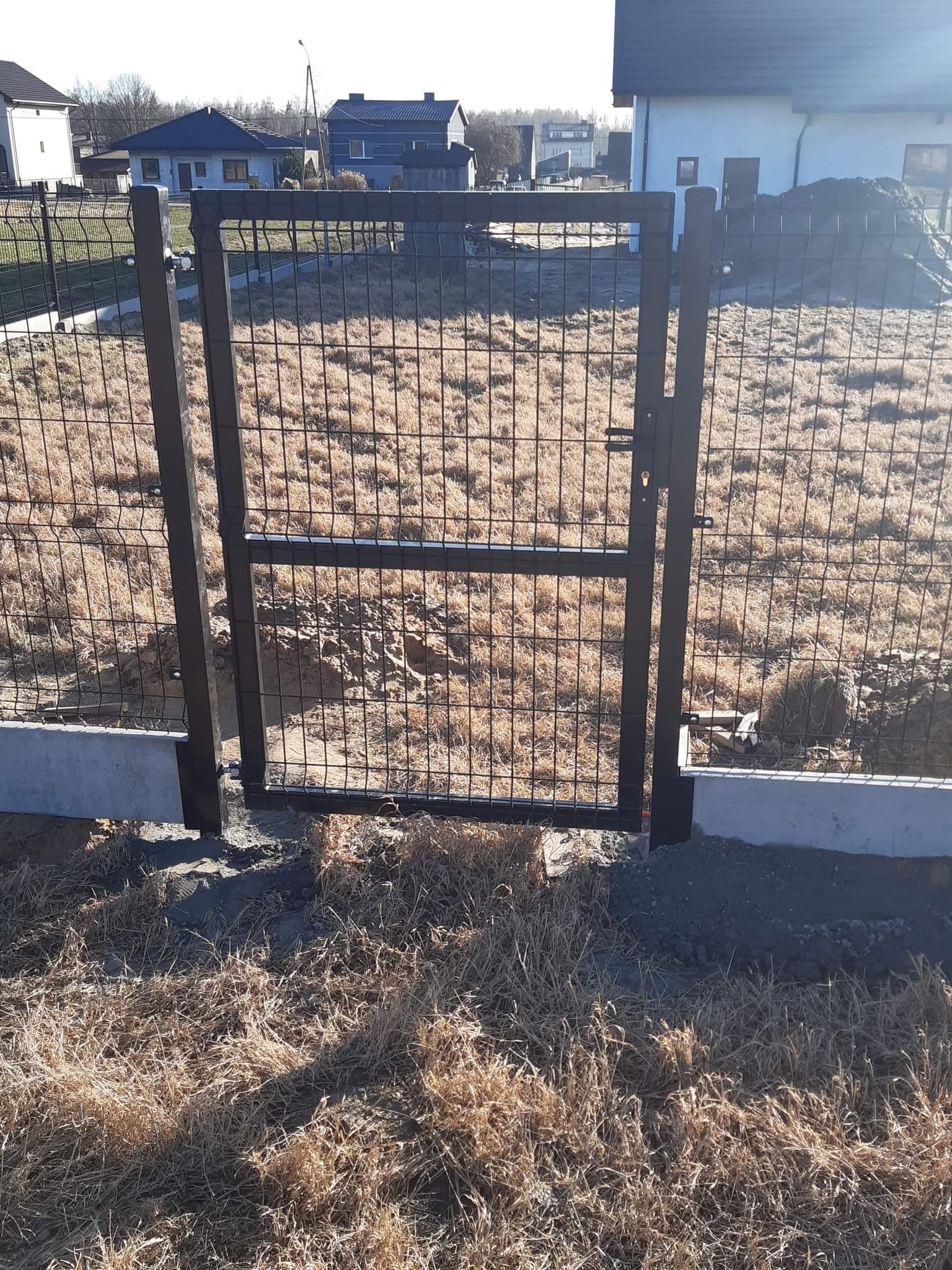 Ciemna brama z siatki ogrodzeniowej zamontowana na betonowej podmurówce, widok na trawnik i domy w tle.