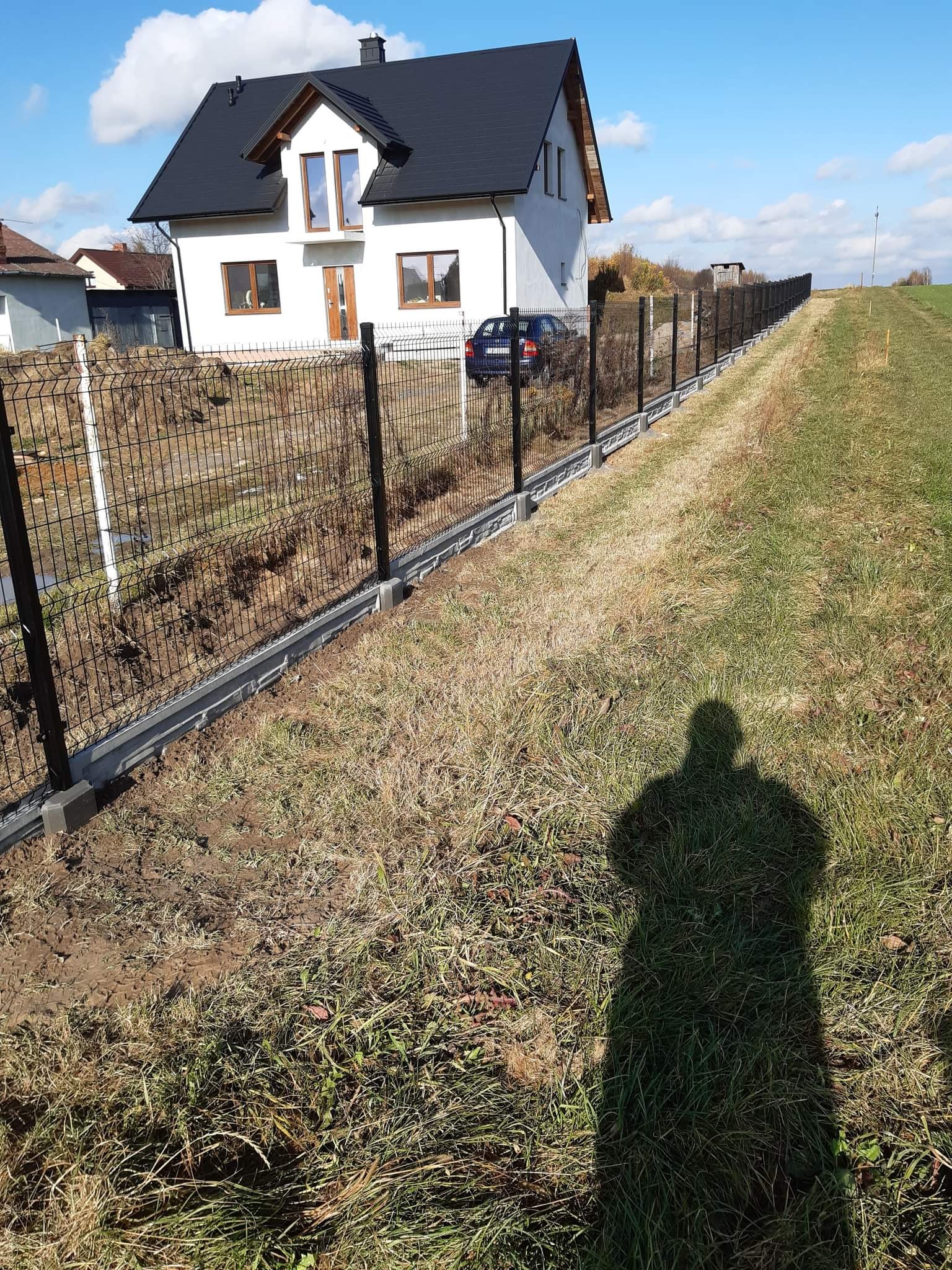 Długa linia ogrodzenia panelowego z podmurówką betonową, biegnąca wzdłuż granicy działki, z widocznym nowym domem w tle i cieniem fotografa na pierwszym planie.