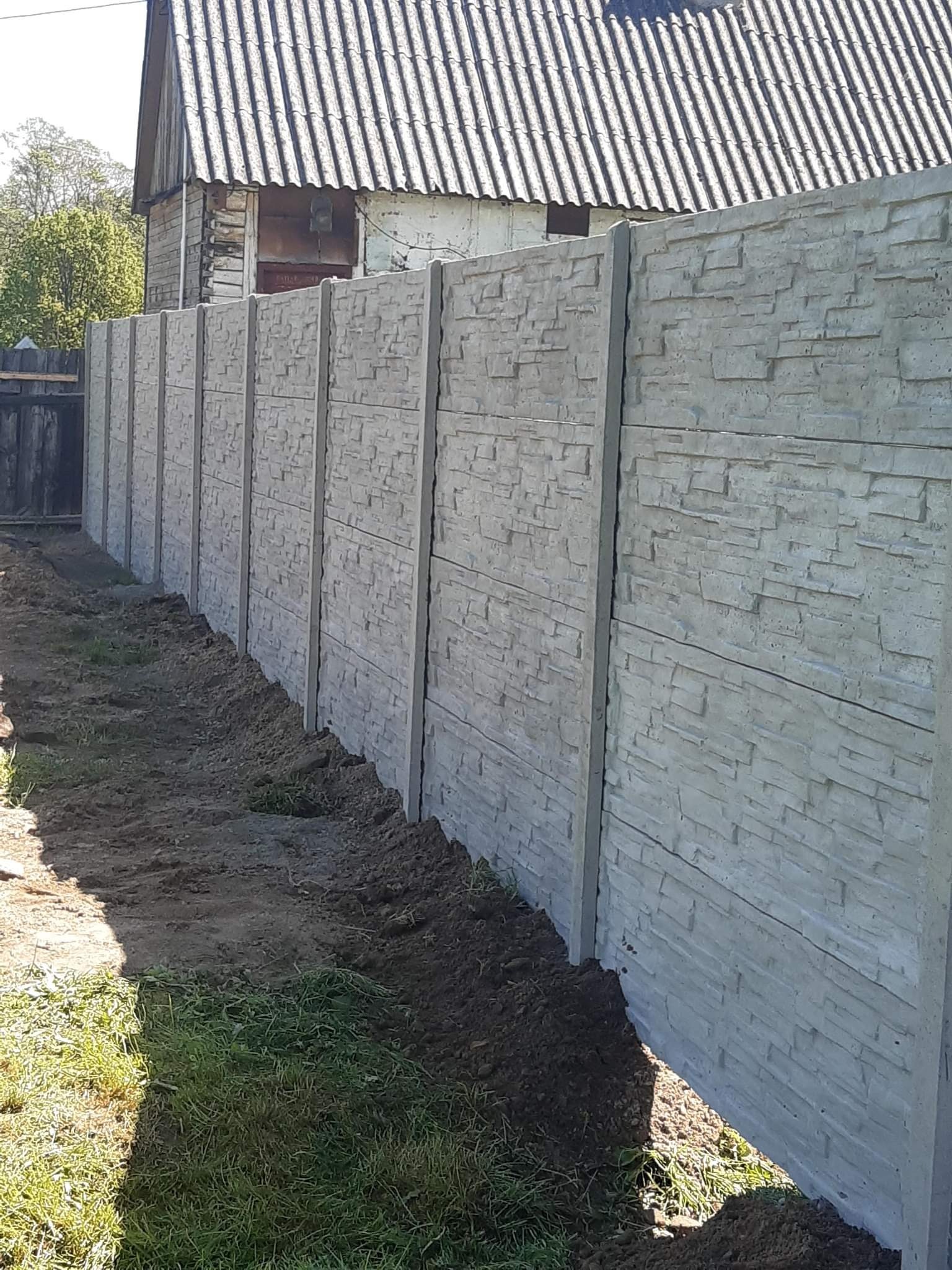 Długi, szary mur z prefabrykowanych elementów betonowych imitujących kamień, oddzielający posesję od drogi, z widocznym domem w tle.