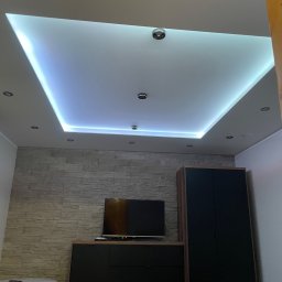 SELMO - Pokój z podwieszanym sufitem z oświetleniem LED, ściana wykończona imitacją kamienia, czarne meble i telewizor.