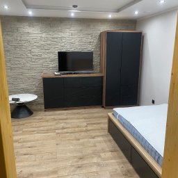 SELMO - Nowoczesna sypialnia z kamienną ścianą, czarnymi meblami z geometrycznym wzorem, drewnianą podłogą i łóżkiem z szarą ramą.