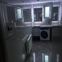 SELMO - Nowoczesna łazienka z szarymi płytkami, podświetlanymi lustrami, pralką pod blatem i grzejnikiem ściennym. Widoczne odbicie osoby w lustrze.