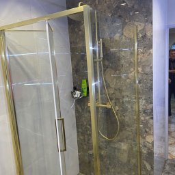 SELMO - Kabina prysznicowa z złotymi profilami i szklanymi drzwiami, widoczna armatura i półka z kosmetykami na tle marmurowych płytek ściennych.