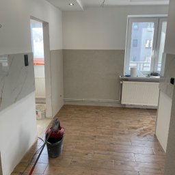 SELMO - Wnętrze w trakcie prac wykończeniowych: brązowe płytki imitujące drewno na podłodze, ściany częściowo wykończone, widoczne narzędzia i wiadro z zaprawą, sufit z oświetleniem punktowym i wystającymi...