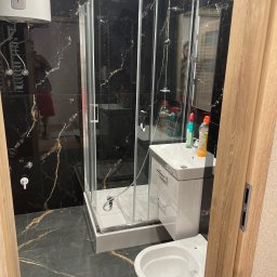 SELMO - Czarno-złota łazienka z kabiną prysznicową, umywalką z szafką i podwieszaną toaletą, widok przez drewniane futryny drzwi.