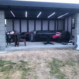 SELMO - Zadaszona altana w trakcie budowy z betonowych bloczków, z surowym betonowym podłożem i prowizorycznym oświetleniem LED, częściowo umeblowana czarną sofą i stołem.