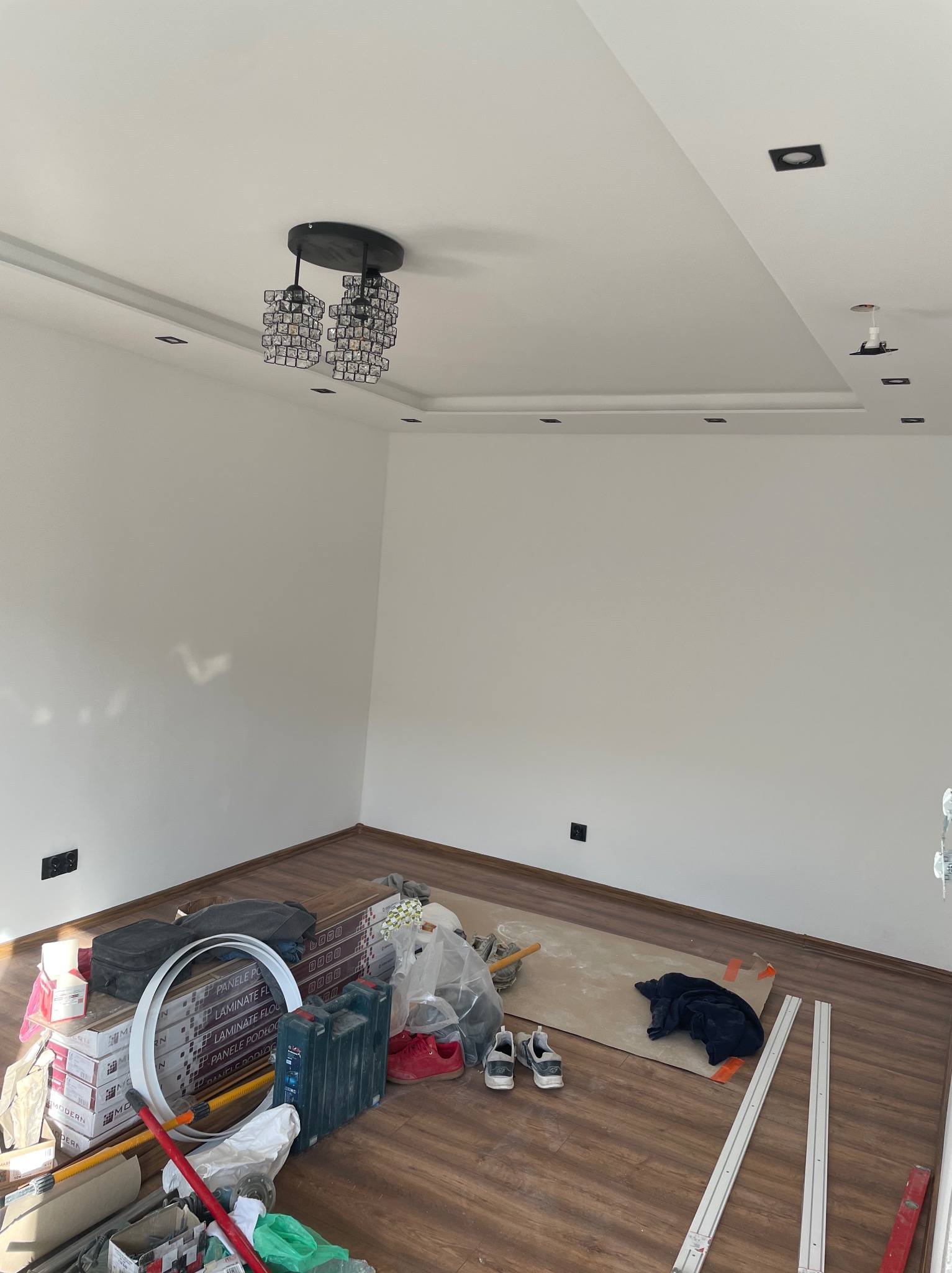 Pokój w trakcie prac wykończeniowych: ułożone panele podłogowe, białe ściany, sufit z zamontowanym nowoczesnym żyrandolem i oświetleniem punktowym, widoczne materiały budowlane i narzędzia.