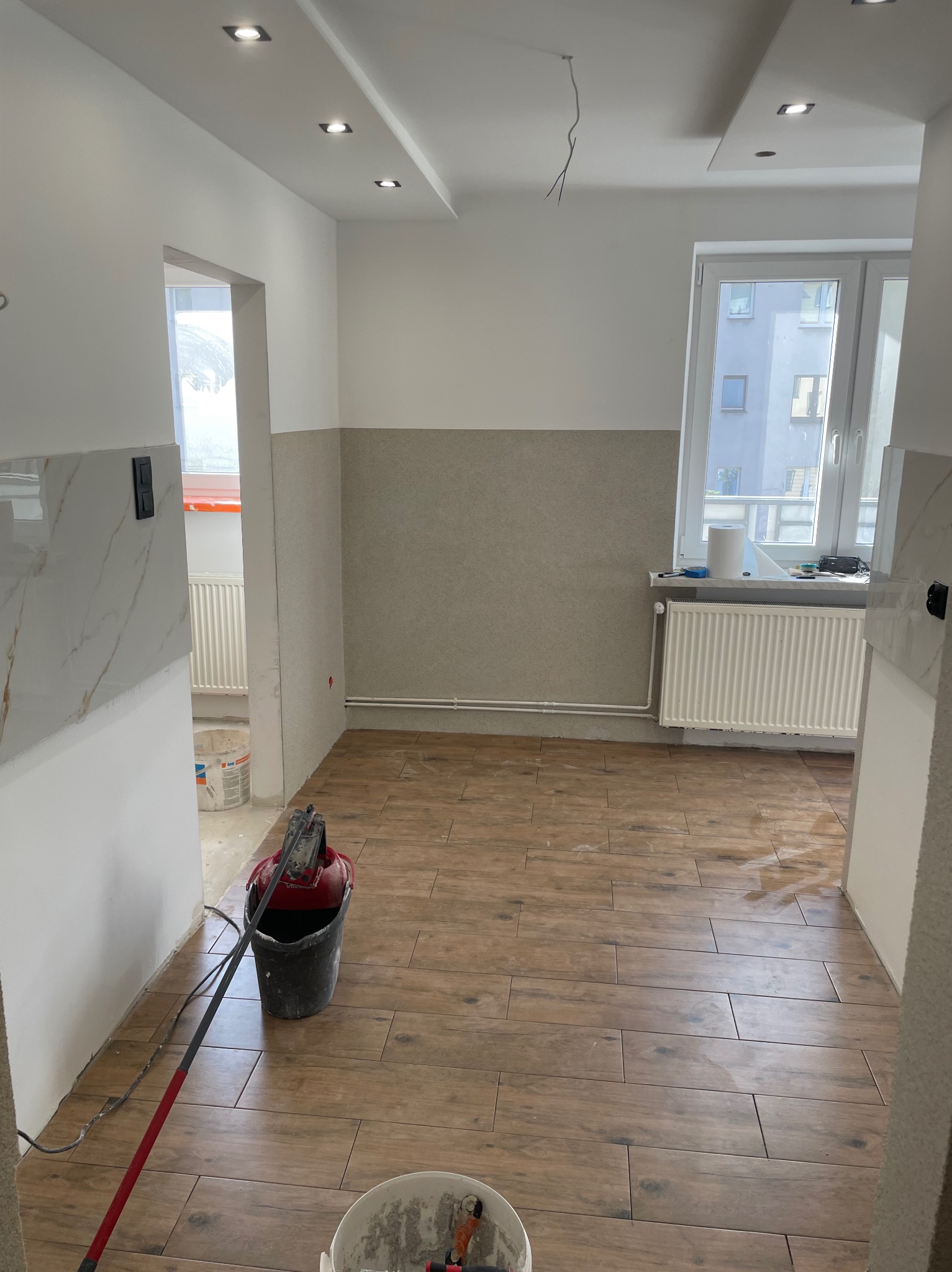 Wnętrze w trakcie prac wykończeniowych: brązowe płytki imitujące drewno na podłodze, ściany częściowo wykończone, widoczne narzędzia i wiadro z zaprawą, sufit z oświetleniem punktowym i wystającymi...