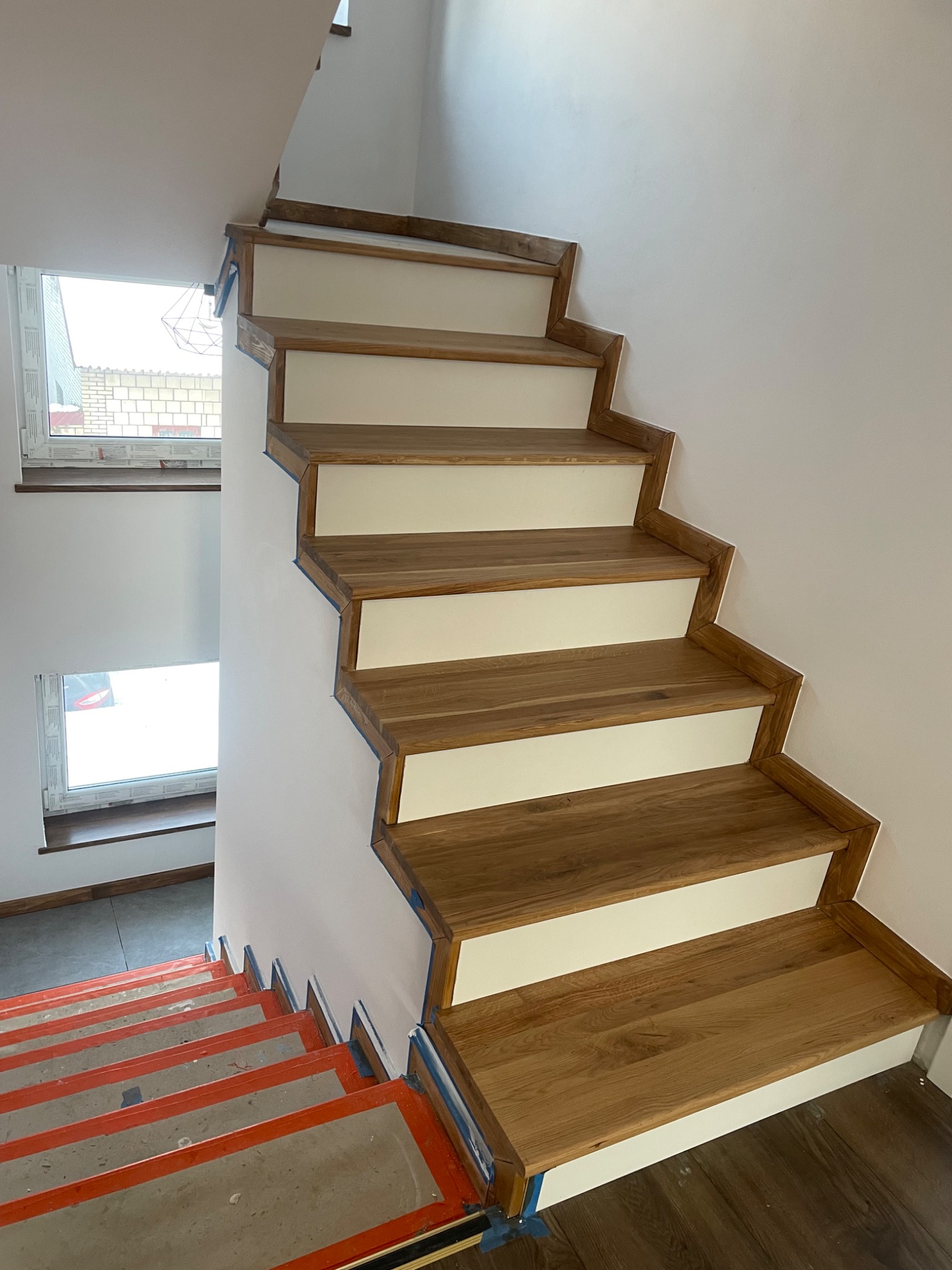 Drewniane schody z białymi podstopnicami w trakcie montażu, zabezpieczone taśmą malarską, widok z góry na dolne stopnie z zabezpieczeniem podłogi.