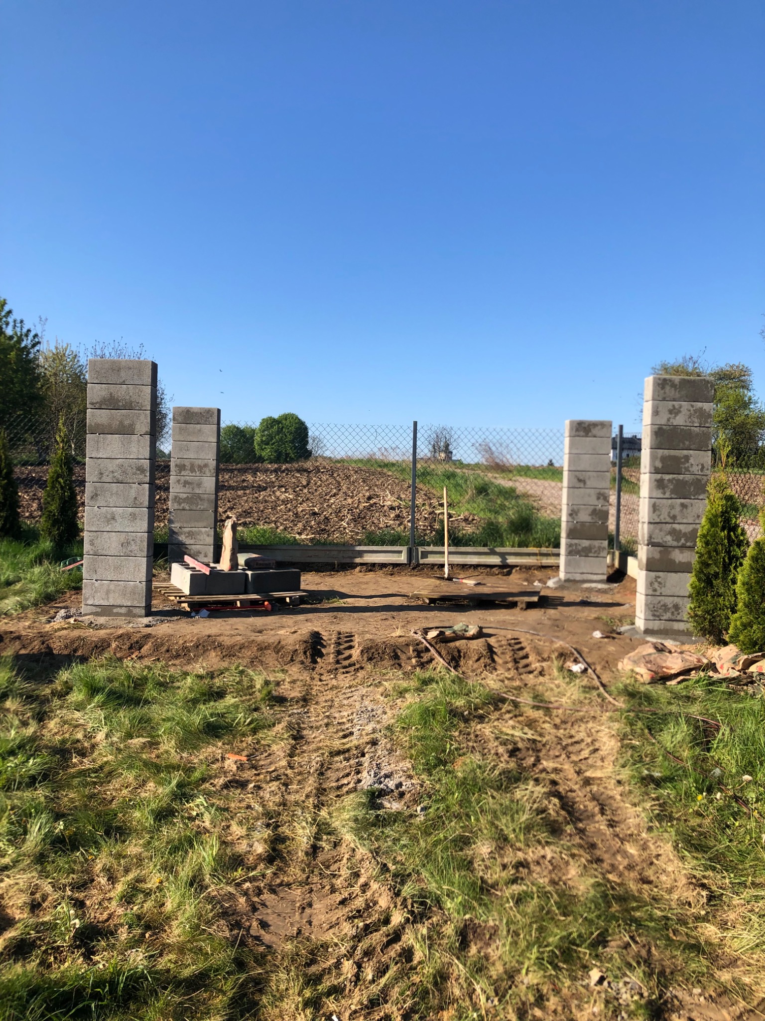 Wznoszenie słupków ogrodzeniowych z bloczków betonowych, widoczne narzędzia i poziomica, teren przygotowany pod budowę ogrodzenia panelowego.
