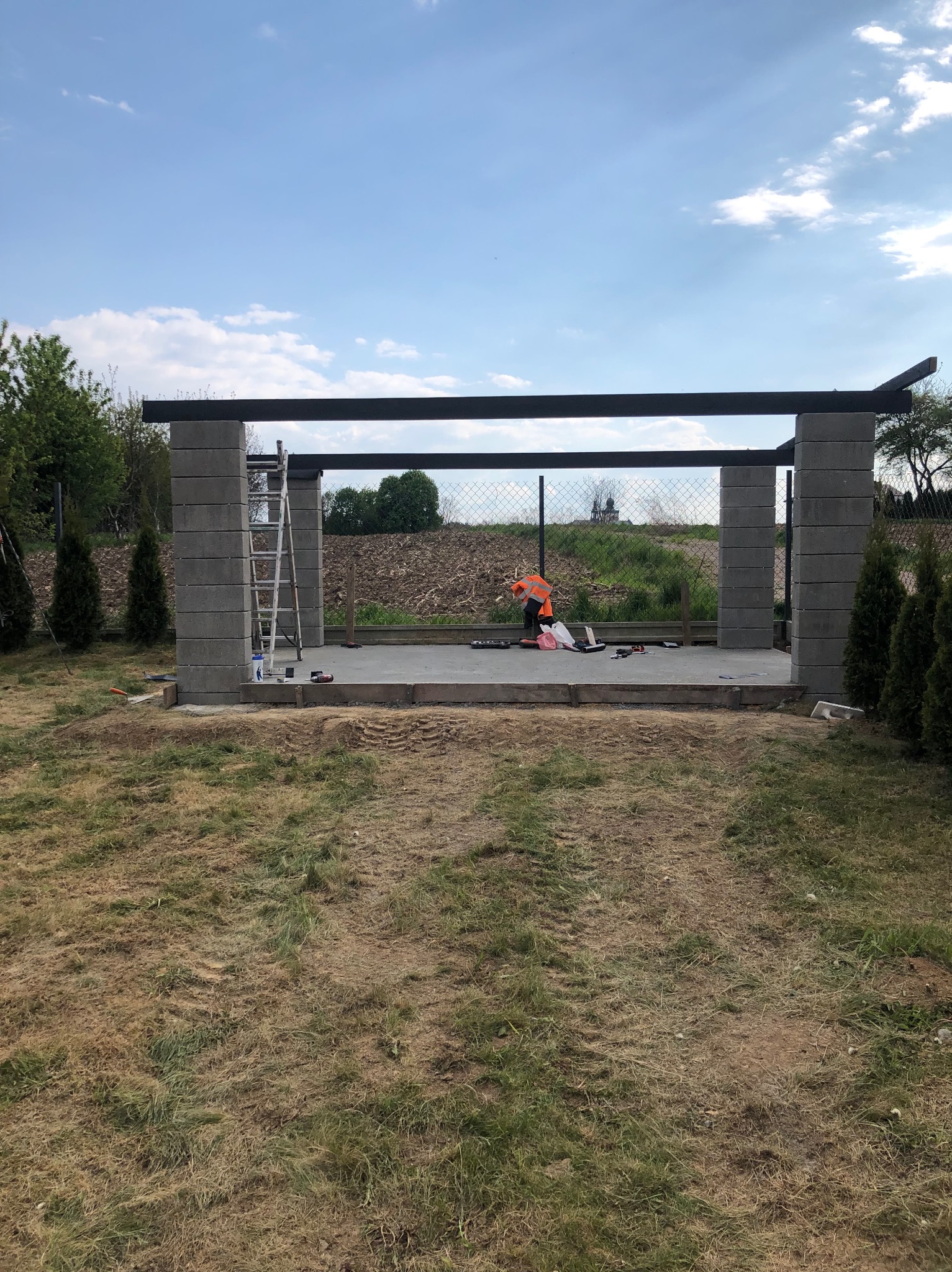Budowa zadaszenia tarasu w trakcie: widoczne betonowe słupy, metalowa konstrukcja dachu, pracownik w kamizelce odblaskowej oraz narzędzia na betonowej wylewce.