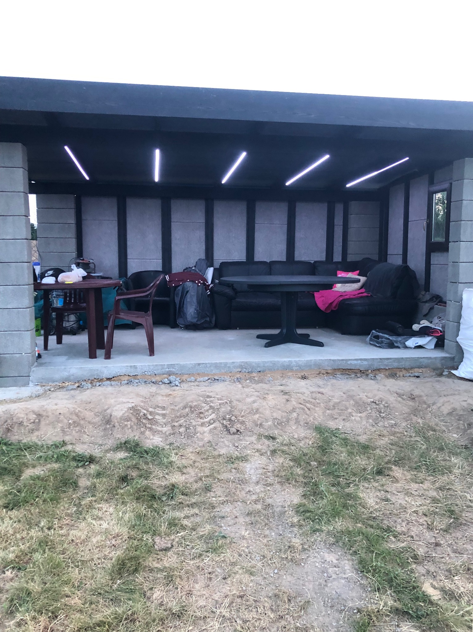 Zadaszona altana w trakcie budowy z betonowych bloczków, z surowym betonowym podłożem i prowizorycznym oświetleniem LED, częściowo umeblowana czarną sofą i stołem.