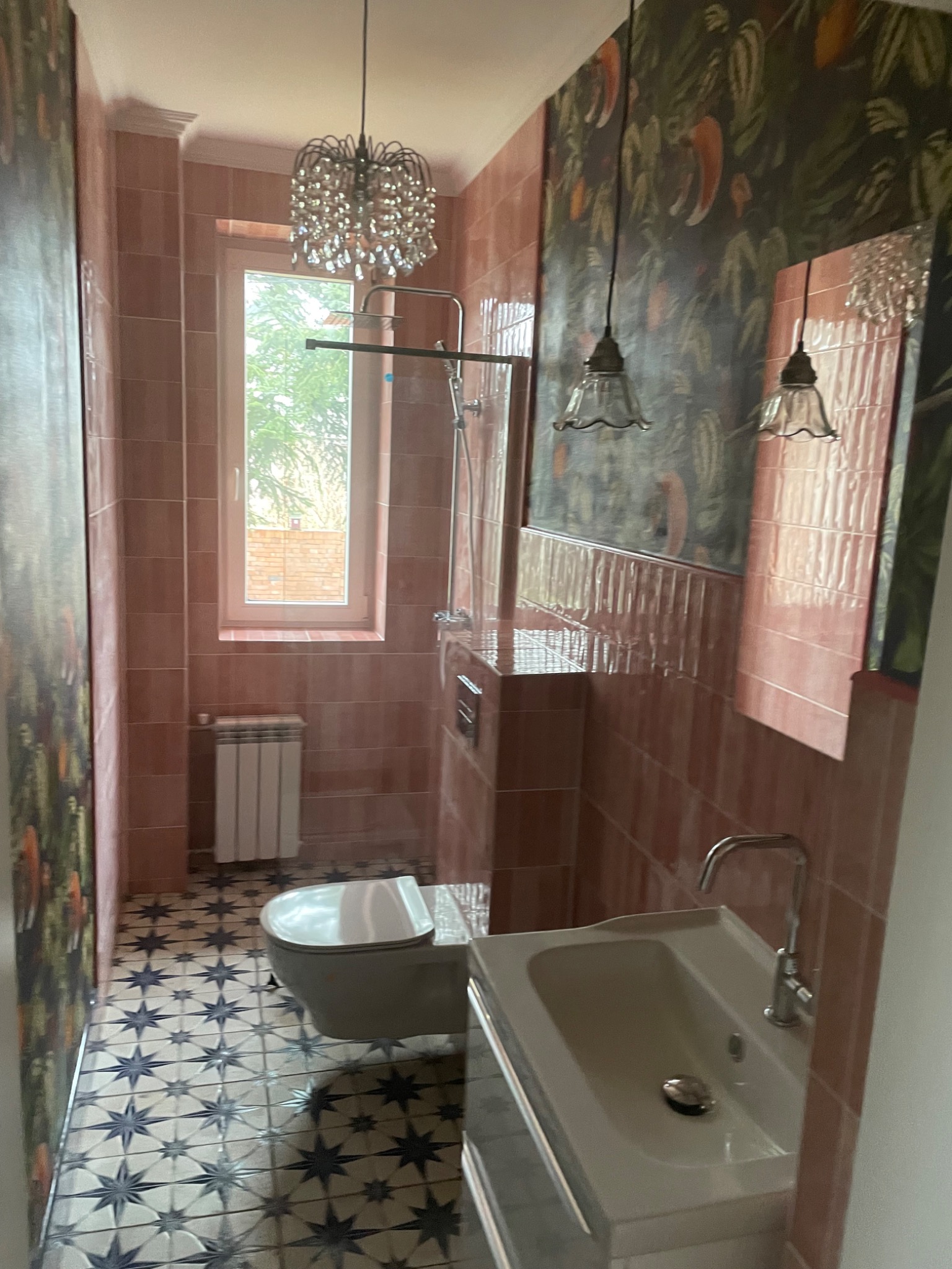 Wykończona łazienka w różowych odcieniach z wzorzystą podłogą, wiszącą toaletą i umywalką. Na ścianach płytki i tapeta w roślinny wzór. Widoczne oświetlenie.