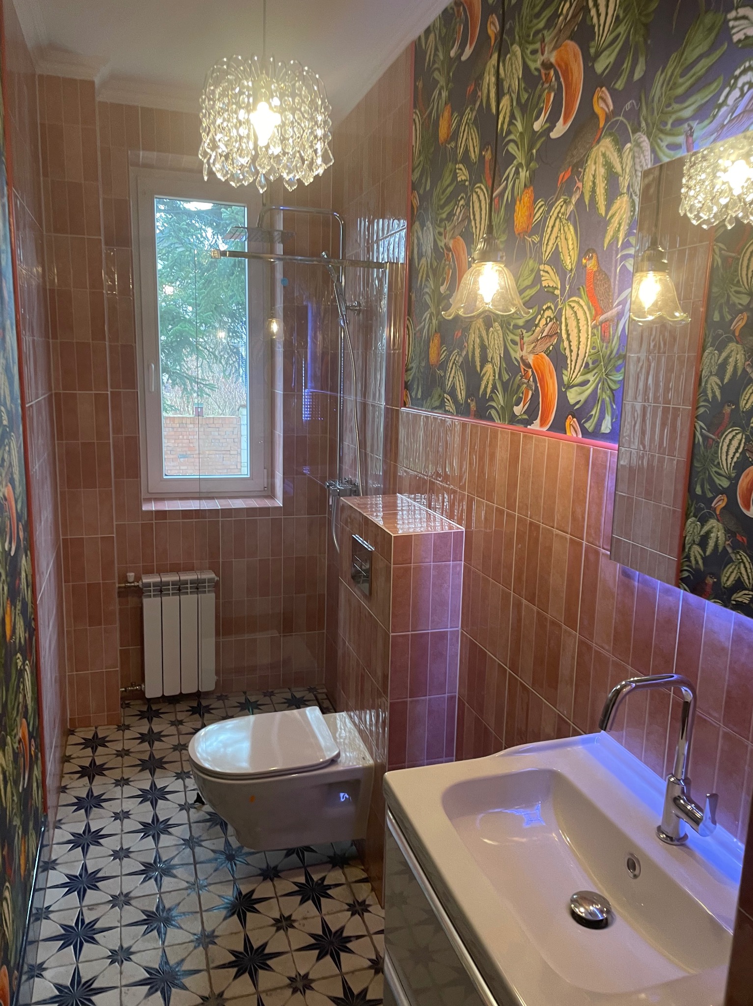 Wykończona łazienka z beżowymi płytkami, wzorzystą tapetą i podłogą w gwiazdy. Widoczna umywalka, toaleta i dekoracyjna lampa sufitowa.