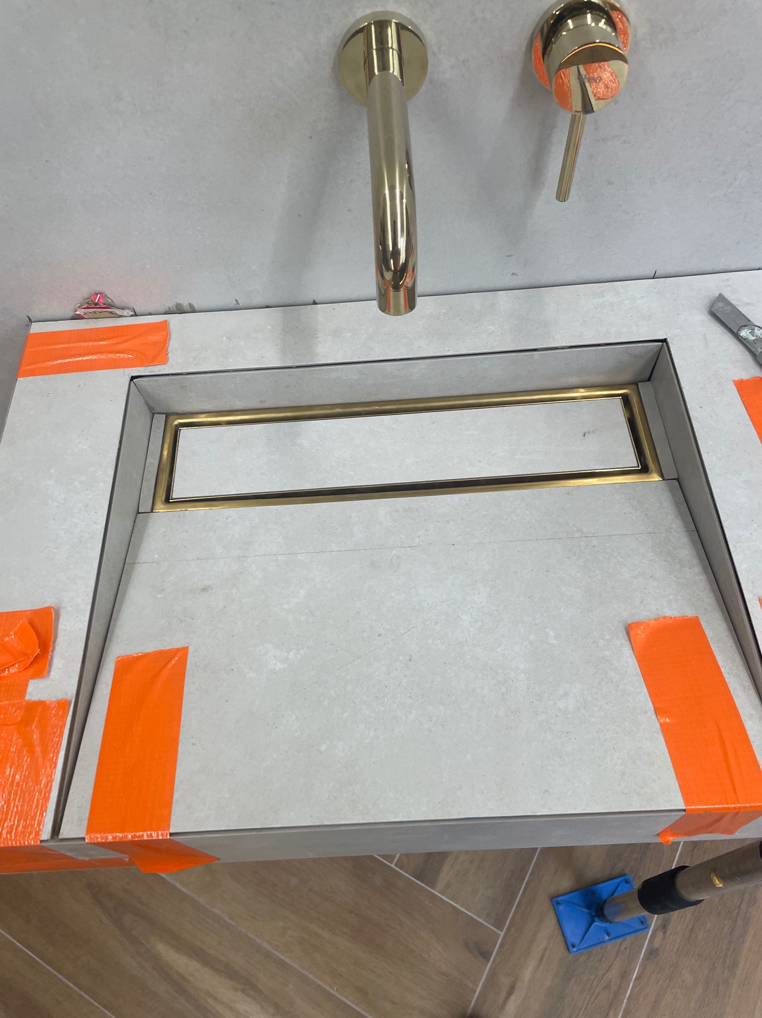 Białe płytki z złotym odpływem liniowym zabezpieczone pomarańczową taśmą, złota bateria umywalkowa, drewnopodobna podłoga. Etap prac wykończeniowych w łazience.