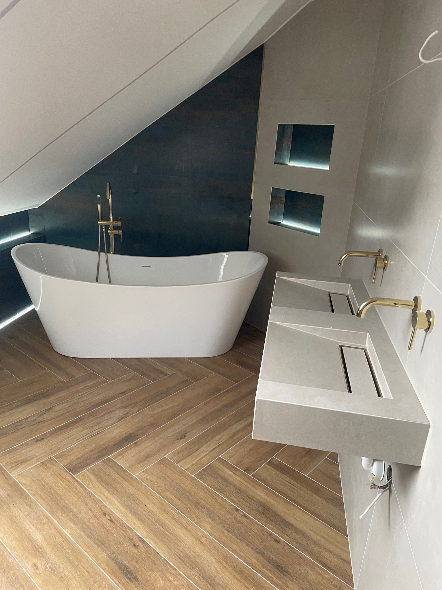 Nowoczesna łazienka na poddaszu: wolnostojąca wanna, podwójna umywalka z złota armaturą, drewnopodobna podłoga w jodełkę i oświetlenie LED.