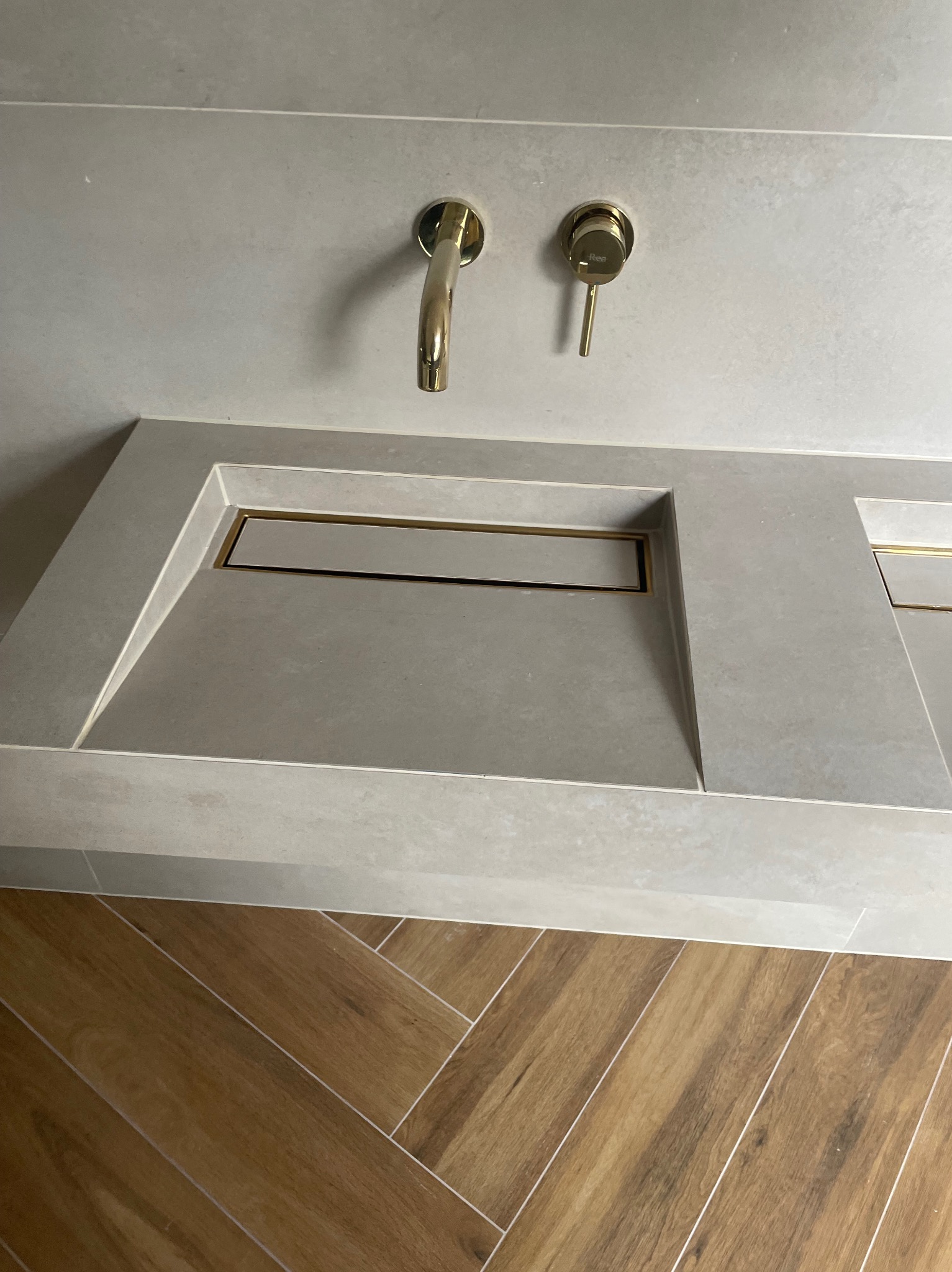 Nowoczesna umywalka z szarego kamienia z złotą armaturą i odpływem liniowym, położona na drewnianej podłodze w jodełkę. Minimalistyczny design.