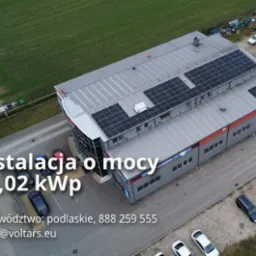 Widok z góry na budynek z zainstalowanymi panelami słonecznymi na dachu, obok parkingu z samochodami, z widoczną informacją o mocy instalacji 30,02 kWp.