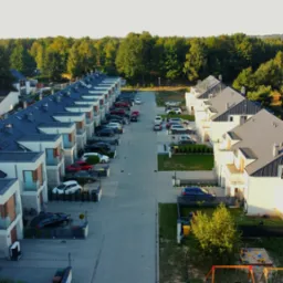 Osiedle dom&oacute;w jednorodzinnych w zabudowie szeregowej widziane z g&oacute;ry, z panelami słonecznymi na dachach, samochodami zaparkowanymi wzdłuż ulicy i placem zabaw w oddali.