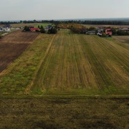 FMdronephotography - Szeroki kadr z lotu ptaka na wiejskie pole po żniwach, z widocznymi śladami po kombajnie, w tle zabudowania wiejskie i las.