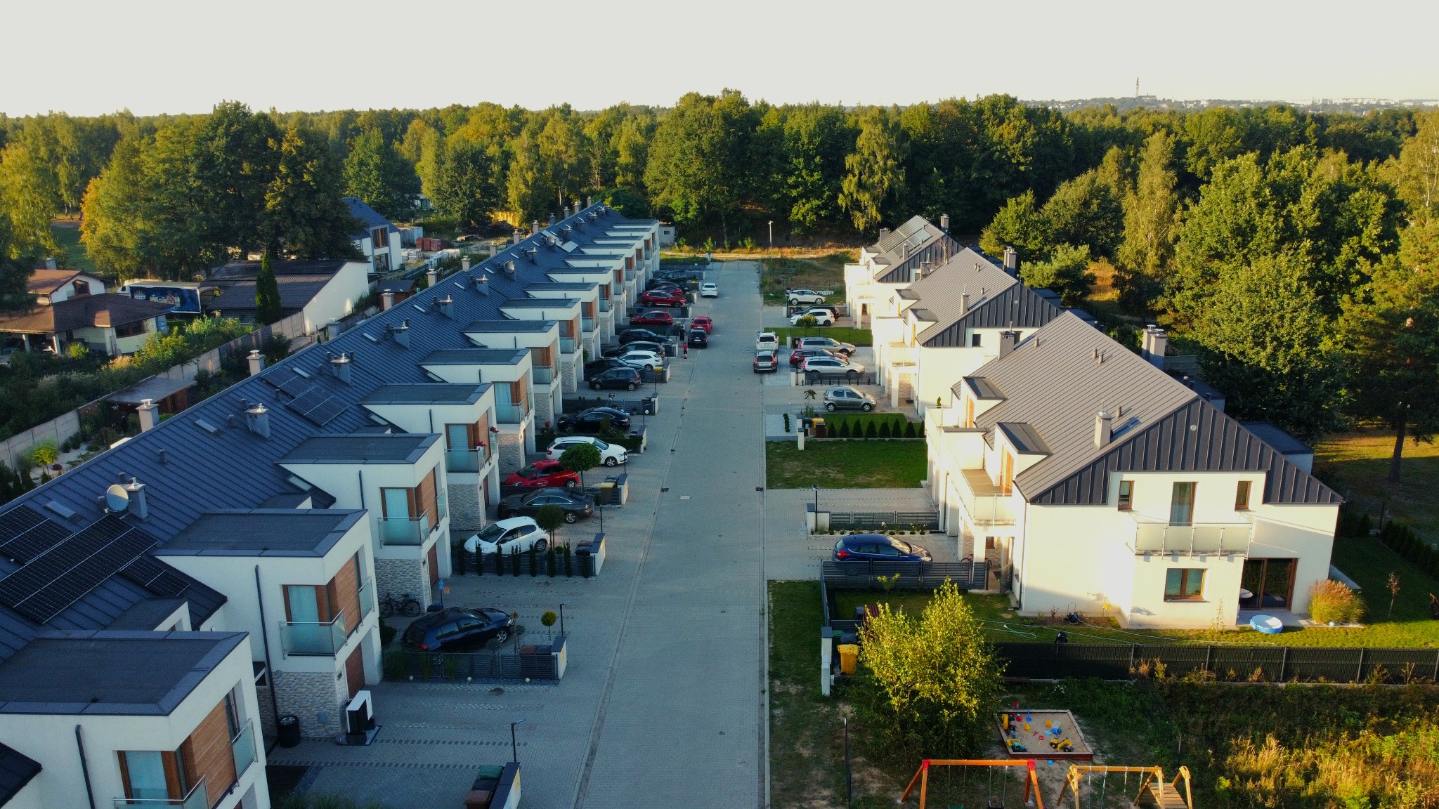 Osiedle domów jednorodzinnych w zabudowie szeregowej widziane z góry, z panelami słonecznymi na dachach, samochodami zaparkowanymi wzdłuż ulicy i placem zabaw w oddali.
