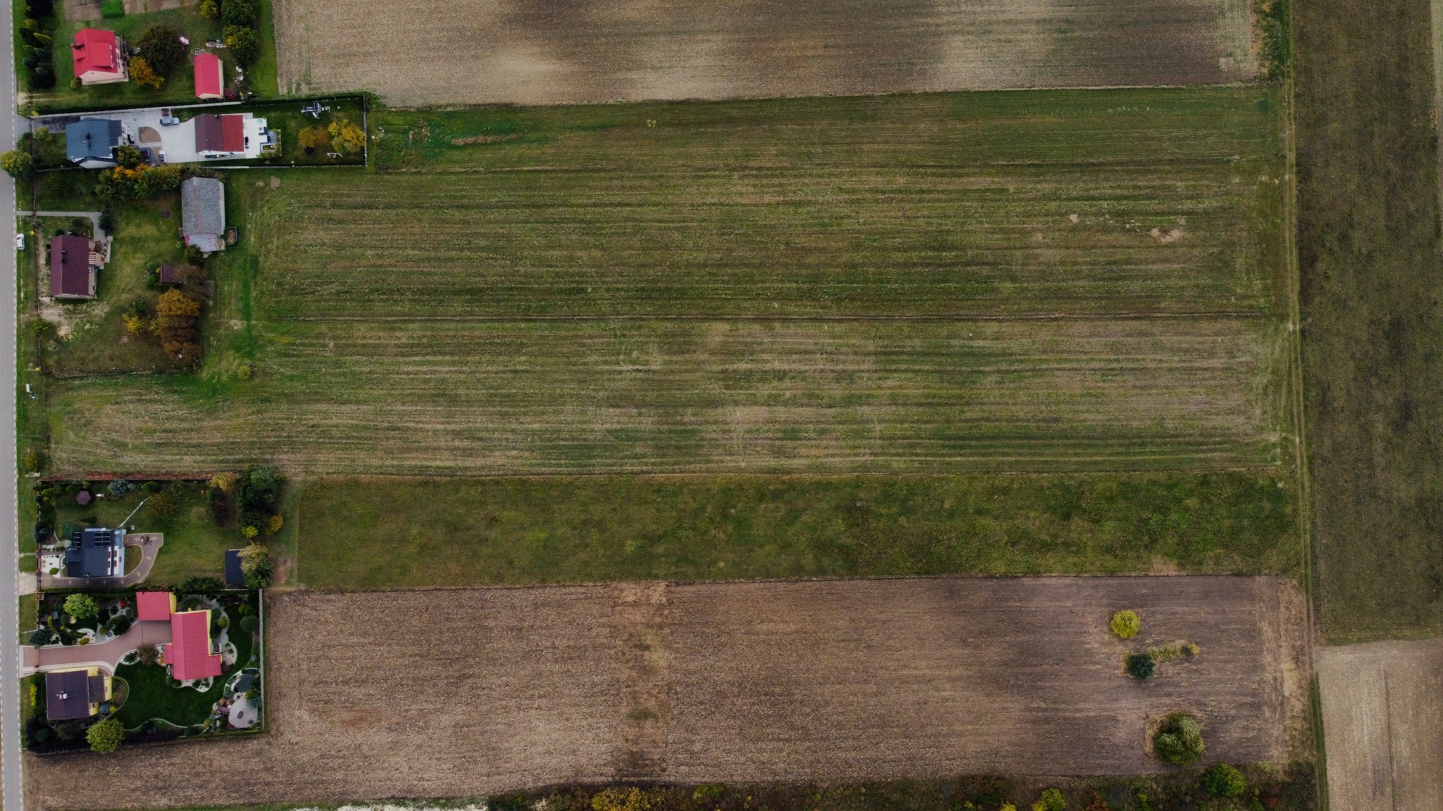 Zdjęcie z drona przedstawiające mozaikę pól uprawnych o różnych odcieniach zieleni i brązu, sąsiadujących z zabudowaniami mieszkalnymi i drogą.