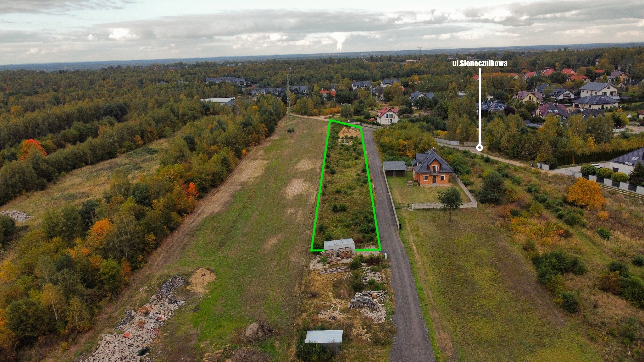 Działka budowlana z naniesionym zielonym obrysem, widok z drona na obrzeżach miejscowości, w tle las i zabudowa jednorodzinna, adres ul. Słonecznikowa.
