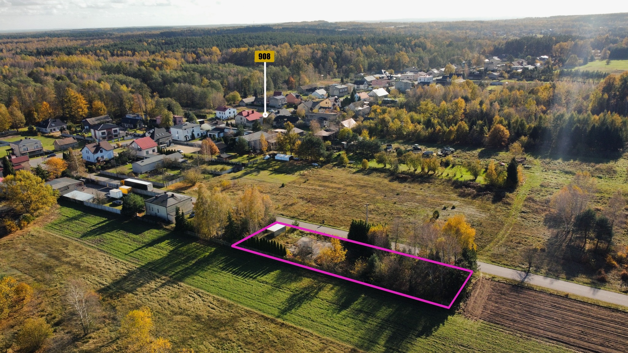 Zdjęcie z drona przedstawiające działkę budowlaną z zaznaczonym obrysem, otoczoną polami i lasem w jesiennej scenerii, z widoczną zabudową wiejską w tle.