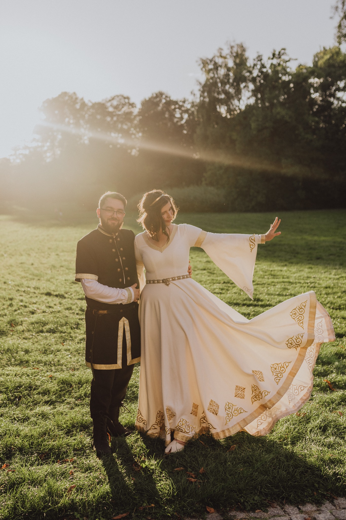 Para w stylizowanych strojach średniowiecznych na tle zieleni i promieni słonecznych. Kobieta w sukni z szerokimi rękawami, mężczyzna w ciemnym kaftanie z ozdobnymi detalami.