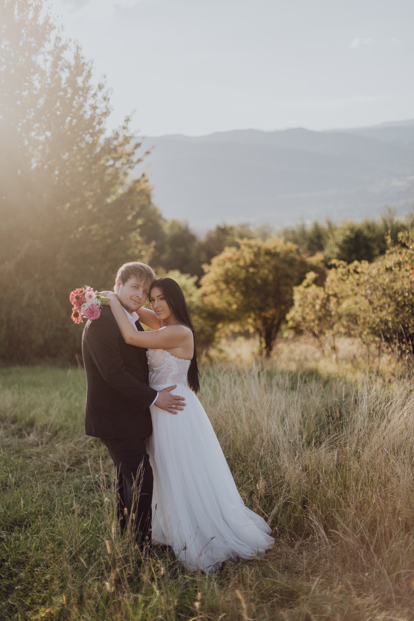 Para młoda w objęciach na tle łąki i gór w słoneczny dzień. Panna młoda w białej sukni, pan młody w czarnym garniturze. Bukiet kwiatów w rękach pana młodego.