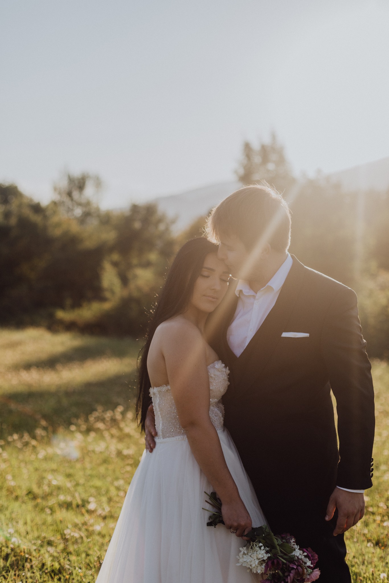 Para młoda w objęciach na łące w promieniach zachodzącego słońca. Panna młoda w sukni z bukietem, pan młody w garniturze. Romantyczna sesja ślubna w plenerze.