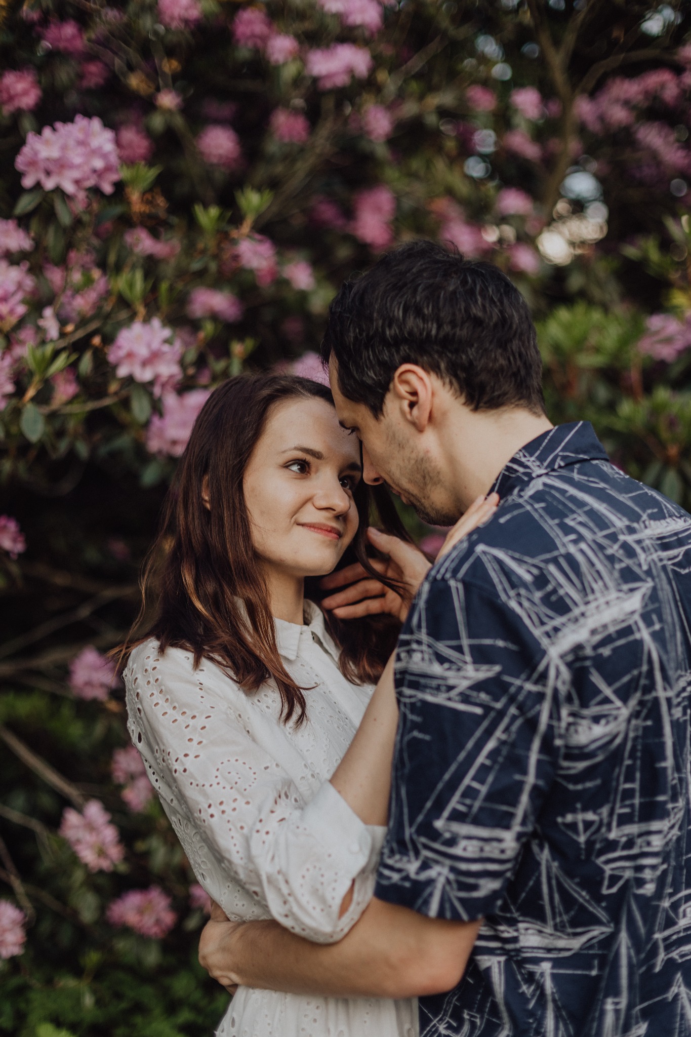Zakochana para przytulona na tle kwitnącego krzewu różowych kwiatów. Kobieta w białej bluzce spogląda z uśmiechem na mężczyznę w koszuli w żaglówki.