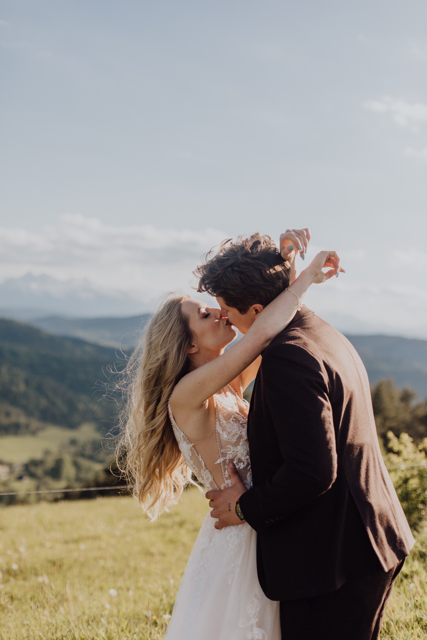 Pocałunek młodej pary na tle gór. Panna młoda w koronkowej sukni, pan młody w brązowym garniturze. Romantyczna sceneria, naturalne światło, delikatne kolory.