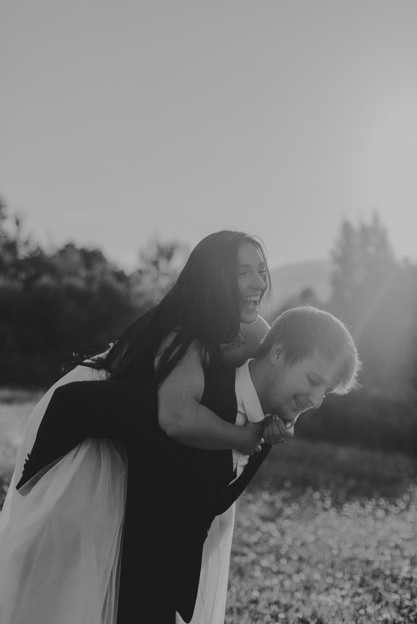 Radosna para młoda w czerni i bieli: panna młoda na barana u pana młodego, oboje śmieją się w promieniach zachodzącego słońca na tle łąki.