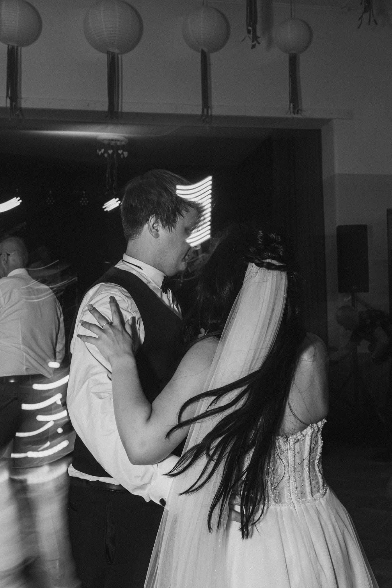 Czarno-białe ujęcie tańczącej pary młodej w Bielsku-Białej. Panna młoda w welonie i sukni, pan młody w kamizelce. W tle dekoracje sali i rozmazane sylwetki gości.